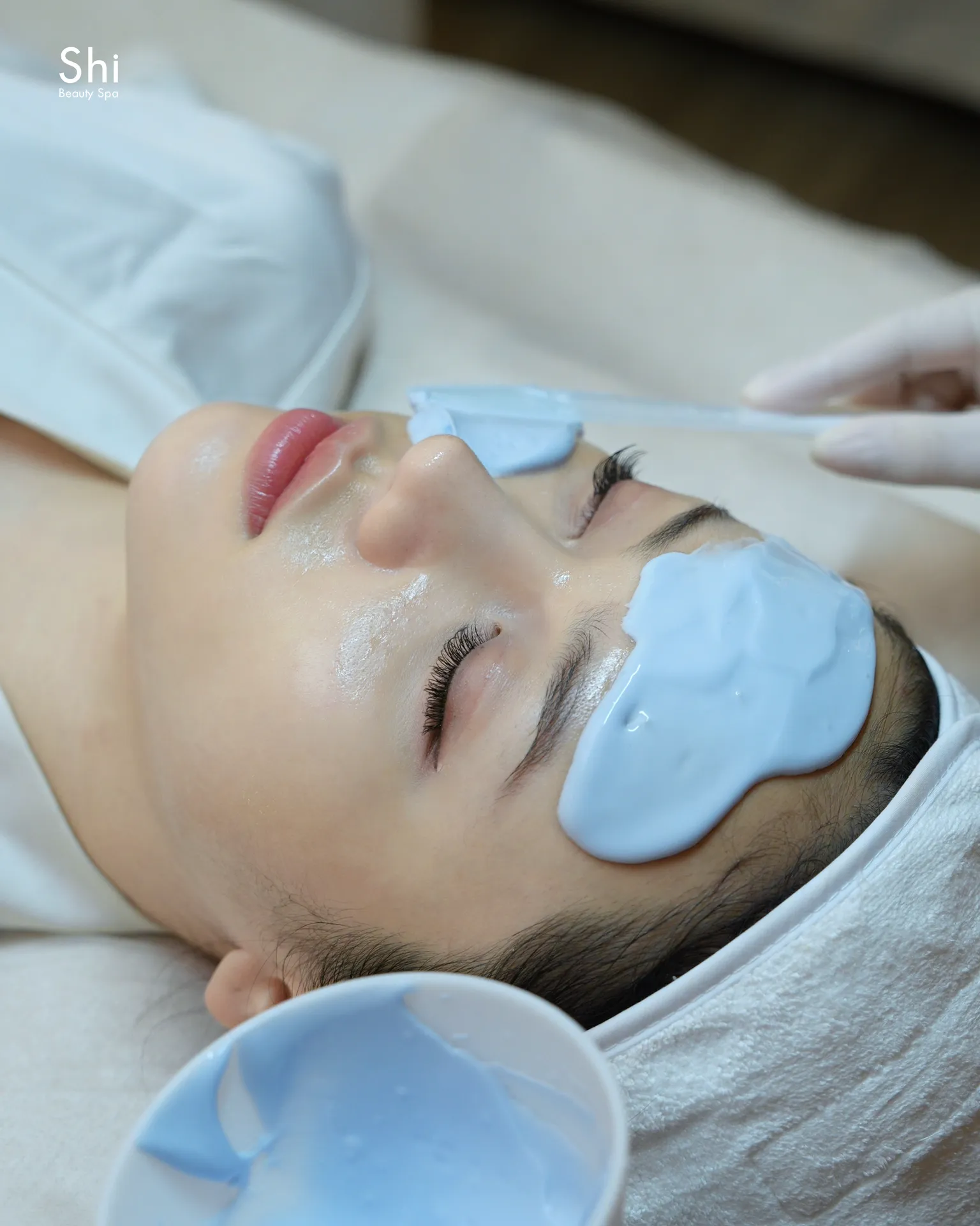 Chuyên viên Shi đeo găng tay dùng cọ đắp lớp mặt nạ lên da mặt khách đang nằm trên giường